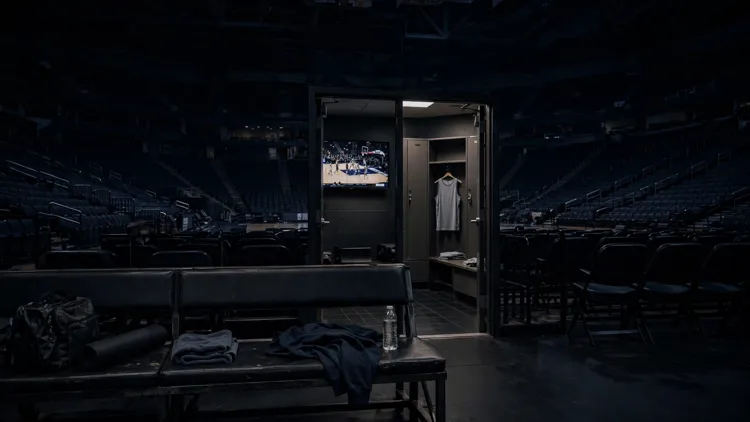 An empty visitor's bench in a darkened basketball arena with a television in a locker room behind it showing a game in progress, a single unbranded jersey hanging nearby, editorial graphic
