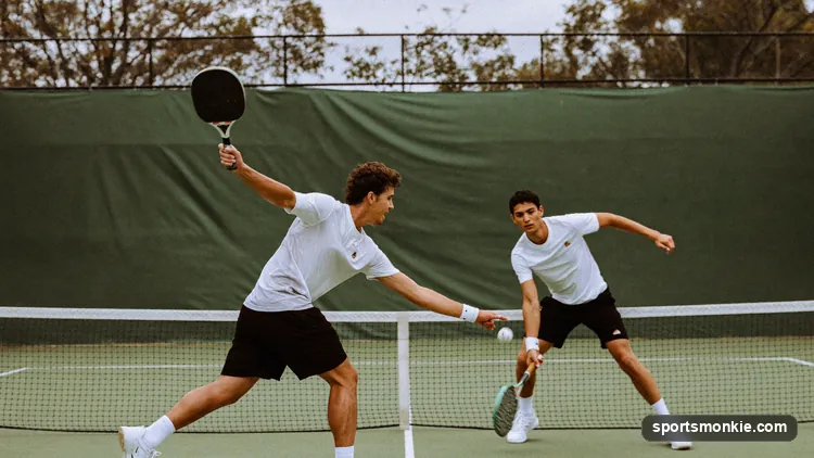 A pickleball doubles rally in progress with the scoreboard visible courtside