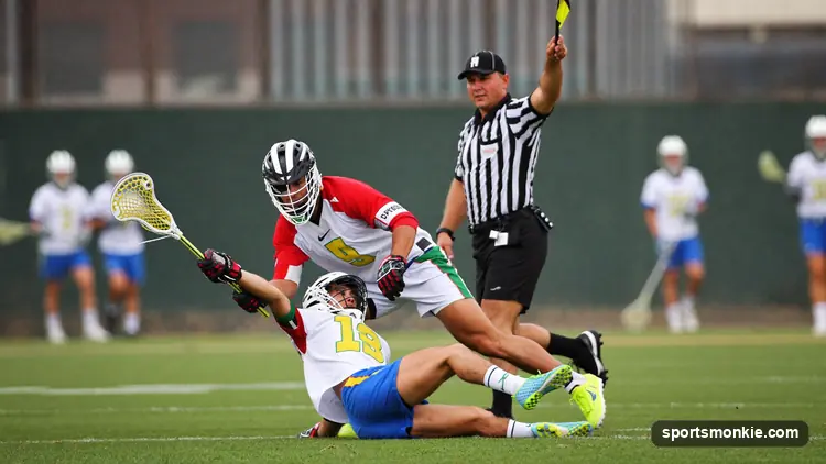 A men's lacrosse defender swinging his stick at an opponent's arm as a referee raises a flag on the field